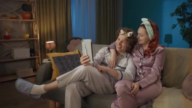Aile ve ilişkiler reklam konsepti. Birlikte zaman geçiren genç bir çiftin portresi. Pijamalı bir adam ve kadın, saç bantlarıyla kanepede oturmuş tabletteki video konuşmalarıyla konuşuyorlar..