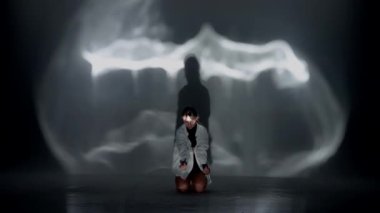 Üstü açık, şortlu ve tişörtlü genç bir kadın stüdyodaki ışığın altında duygusal anlamda çağdaş dans ediyor. Arka planda siyah bir gölge var. Tam uzunluk.