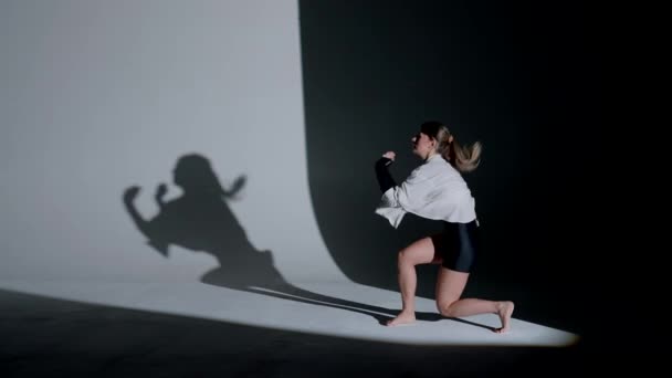 トップ ショートパンツ スタジオでスポットライトの下で感情的な現代舞踊を行うシャツを着ている若い女性 背景に黒い落下影をコントラストする 全長について — ストック動画