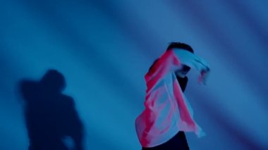 Stüdyoda üstsüz, şortlu ve tişörtlü genç bir kadın duygusal dans gösterisi yapıyor. Neon mavi ve kırmızı renk şeması, gölgeli arka plan. Tam boy. Reklam, yaratıcı içerik.