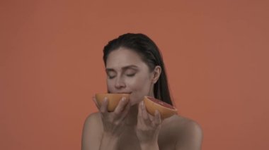 Güzel esmer modelin portresi. Doğal makyajlı ve gülümseyen bir kadını yakından çek. Cilt bakımı. Greyfurt yarımları tutan, gülümseyen ve ısıran çekici bir kadın. Yavaş çekim