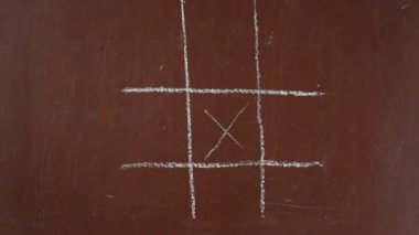 Kalıplı kahverengi karatahta arka planı. Erkek el bir tic-tac-toe oynuyor, isim veriyor ve yakın planda bir parça beyaz tebeşirle oyunu geçiyor. Eğitici ve yaratıcı içerik, okul kavramı.