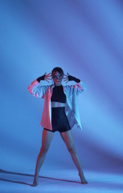 Stüdyoda üstsüz, şortlu ve tişörtlü genç bir kadın duygusal dans gösterisi yapıyor. Neon mavi ve kırmızı renk şeması, gölgeli arka plan. Tam boy. Reklam, yaratıcı içerik.