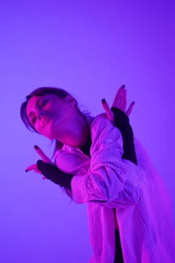 Stüdyoda üstsüz, şortlu ve tişörtlü genç bir kadın modern dans gösterisi yapıyor. Neon pembe ve mor renk şeması, ombre, eğim arkaplanı. Orta boy..