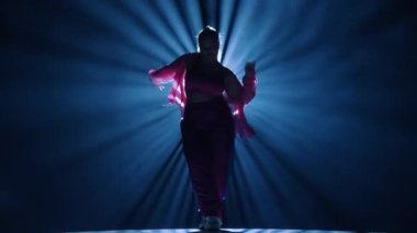 Stüdyoda dans eden bir kadının silueti. Siyah arka plan dumanla dolu, beyaz spot ışıkları arkadan parlıyor. Modern koreografi. Tam boy. Reklam videosu veya reklamı.