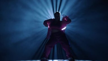 Stüdyoda dans eden bir kadının silueti. Siyah arka plan dumanla dolu, beyaz spot ışıkları arkadan parlıyor. Modern koreografi. Tam boy. Reklam videosu veya reklamı.