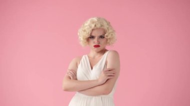 Genç bir kadın, dudaklarını kırmızı rujla büzüyor ve kollarını göğsünde kavuşturuyor. Marilyn Monroe suretinde beyaz bir elbise ve beyaz bir peruk takmış bir kadın portresi.