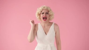 Kadın kafasına beyaz peruk takıyor ve agresif bir şekilde konuşmaya başlıyor, bağırıyor, seni işaret ediyor, suçluyor. Stüdyoda Marilyn Monroe 'nun pembe arka planında kızgın ve üzgün bir kadın var.