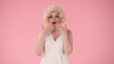 Kadın elleriyle ağzını kapatıyor, işaret parmaklarıyla işaret ediyor, onaylayarak başını sallıyor, tavsiye veriyor. Beyaz elbiseli ve peruklu bir kadın, Marilyn Monroe 'nun stüdyosundaki pembe elbiseli hali.
