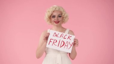 Elinde Kara Cuma yazılı bir poster tutan bir kadın. Pembe arka planda stüdyodaki Marilyn Monroe 'ya benzeyen kadın. İndirim, Kara Cuma, indirimler.