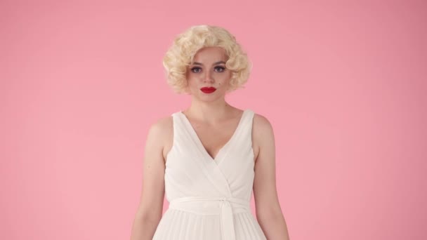 一个年轻女子的画像 天真地微笑着眨了眨眼 玛丽莲 梦露形象中的女人 穿着鲜艳的白衣 戴着粉色背景的假发 — 图库视频影像