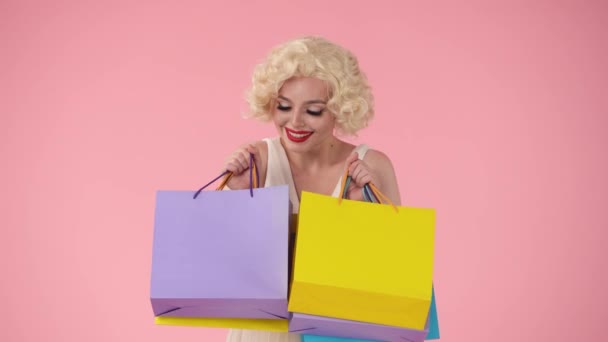 カラフルなショッピングバッグを持っている若い幸せな女性 ピンクのバックグラウンドでスタジオでマリリン モンローのように見える女性 ショッピングコンセプト セールス ブラックフライデー — ストック動画