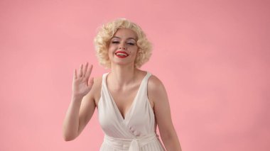 Marilyn Monroe gibi giyinmiş bir kadın selamlaşmak için elini sallıyor. Beyaz elbiseli, beyaz peruklu, dudaklarında kırmızı ruj olan bir kadın stüdyoda, pembe arka planda.