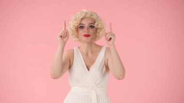 Kadın işaret parmaklarıyla yukarı bakıyor. Beyaz elbiseli ve peruklu bir kadın, Marilyn Monroe 'nun pembe arka plandaki stüdyosuna benziyor. Reklam, tanıtım, kopyalama alanı