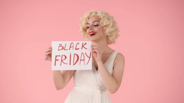 Elinde Kara Cuma yazılı bir poster tutan bir kadın. Pembe arka planda stüdyodaki Marilyn Monroe 'ya benzeyen kadın. İndirim, Kara Cuma, indirimler.