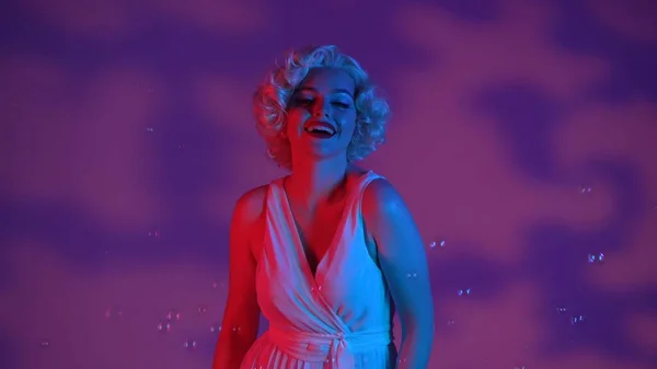Portrait of a cheerful woman looking like Marilyn Monroe surrounded by soap bubbles close up. Woman in studio with highlights, illuminated with pink and green neon light