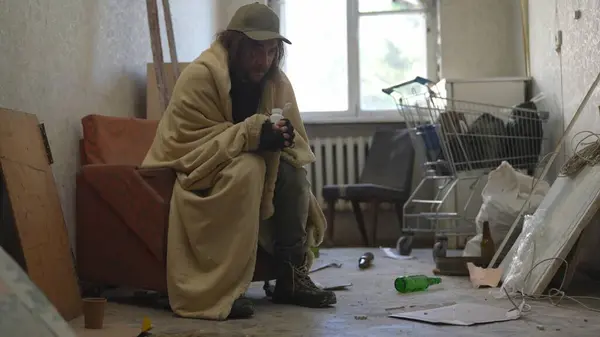 ホームレスの貧しい男は 彼の所有物でいっぱいの放棄された建物の部屋に座っている毛布に覆われた 彼は丸薬のボトルを持っている ホームレスと貧困 — ストック写真