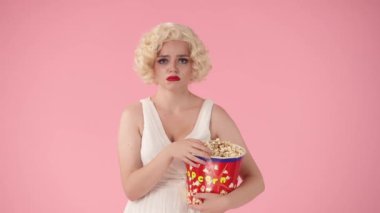 Büyük bir kovadan patlamış mısır yiyen ve film izlerken ağlayan duygusal bir kadının portresi. Pembe arka planda Marilyn Monroe 'ya benzeyen bir kadın.