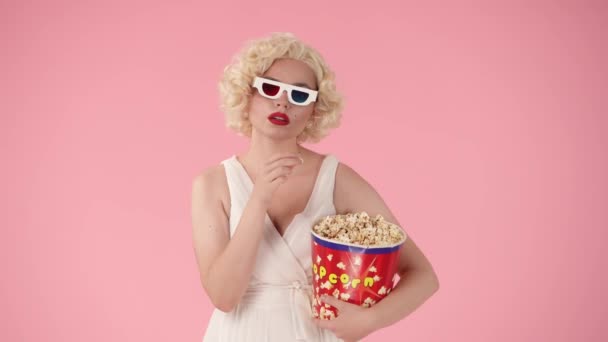 3Dメガネを着て カメラを見て ポップコーンを食べて 怖がって驚きました ピンクの背景にスタジオでマリリン モンローのように見える女性 — ストック動画