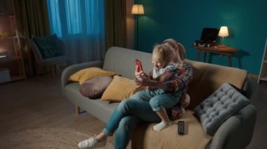 Mutlu anne ve kız, evdeki oturma odasında kanepede otururken akıllı telefondan selfie çekiyorlar. Sevimli küçük kız annesinin kollarında oturuyor ve komik suratlar yapıyor.