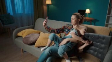 Mutlu anne ve kız, evdeki oturma odasında kanepede otururken akıllı telefondan selfie çekiyorlar. Sevimli küçük kız sevgiyle annesine sarılıyor ve annesi de kızını öpüyor.