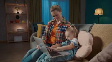 Gülümseyen genç anne küçük kızına bilgisayar uygulamalarını nasıl kullanacağını öğretiyor. Anne ve kızı oturma odasında kanepede oturup online oyun oynuyorlar, çocuklar için eğitim uygulamaları öğreniyorlar, daktilo yazıyorlar.