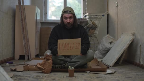 一个无家可归的可怜人坐在一座废弃建筑的房间里 手里拿着一个钱罐 他拿着一块纸板 上面写着无家可归的人 无家可归和贫穷 — 图库视频影像