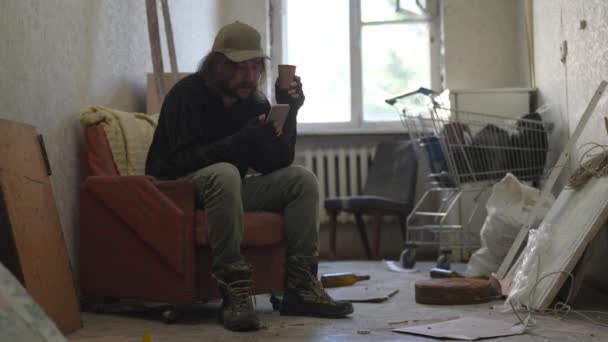 一个无家可归的可怜人坐在一座废弃建筑的房间里 他拿着智能手机 敲打和滚动屏幕 喝着纸杯 无家可归和贫穷 — 图库视频影像