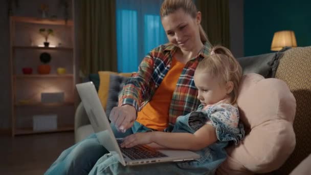 一个母亲教她的小女儿如何使用电脑应用程序的侧视图 妈妈和女儿坐在客厅的沙发上玩网络游戏 为孩子们学习教育应用 — 图库视频影像