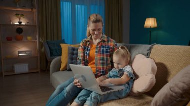 Gülümseyen genç anne küçük kızına bilgisayar uygulamalarını nasıl kullanacağını öğretiyor. Anne ve kızı oturma odasında kanepede oturup online oyun oynuyorlar, çocuklar için eğitim uygulamaları öğreniyorlar, daktilo yazıyorlar.
