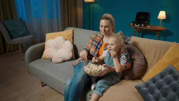 Happy mom sitting on the living room couch cuddled up with her little daughter watching TV and eating popcorn from a big plate close up. Mom and daughter laughing, watching a funny movie, cartoon or