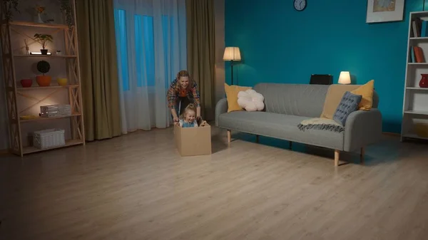 Mom and little daughter having fun. Mom is pushing a cardboard box on the floor, in which the little girl is sitting