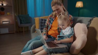 Küçük kızına bilgisayar uygulamalarını nasıl kullanacağını öğreten bir annenin yan görüntüsü. Anne ve kızı oturma odasında kanepede oturup oyun oynuyorlar, çocuklar için eğitim uygulamaları öğreniyorlar.
