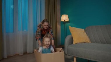 Anne ve küçük kızı eğleniyor. Annem yere bir karton kutu itiyor, küçük kızın oturduğu kutu, yakın çekim. Yavaş çekim