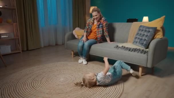 ママと小さな娘は夜のリビングで楽しんでいました 小さなアクティブな女の子が床に振りかけていて お母さんはソファーで座って笑っている — ストック動画