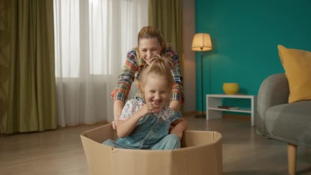 ママと小さな娘が楽しんでいる 母は床にダンボール箱を押し込み 女の子が座ってロリポップを吸い 閉じます スローモーション — ストック動画