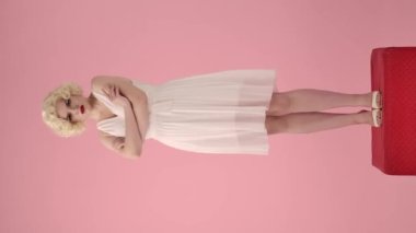 Genç bir kadın gücenmiş duruyor, dudaklarını büzüyor ve kollarını göğsünde kavuşturuyor. Marilyn Monroe 'nun portresindeki bir kadının tam boy portresi. Pembe arka plandaki bir stüdyoda kırmızı bir heykel üzerinde.