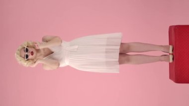 Stüdyoda pembe arka planda kedi gözü güneş gözlüğü takan Marilyn Monroe 'nun portresi. Kırmızı kaidede dikilip beyaz peruk takan kadın poz veriyor. Dikey video
