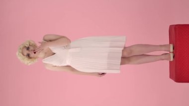 Şaşırmış bir kadın eliyle ağzını kapatıyor, elini işaret ediyor, işaret parmağı yana doğru eğiliyor, onaylayarak başını sallıyor. Marilyn Monroe gibi bir kadın pembe stüdyoda kırmızı kaidede dimdik ayakta duruyor.