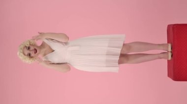 Kadın elleriyle ağzını kapatıyor, işaret parmaklarını yan tarafa doğrultuyor, onaylayarak başını sallıyor, tavsiye veriyor. Marilyn Monroe gibi bir kadın pembe stüdyoda kaidede dimdik ayakta duruyor.