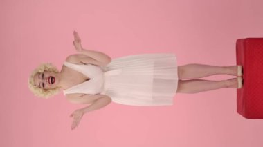 Genç bir kadın zaferini kutlamak için heyecanla zafer jesti yapıyor. Marilyn Monroe 'nun görünümünde bir kadın pembe arka planda kırmızı bir kaidede dikiliyor. Dikey video