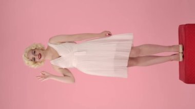 Marilyn Monroe 'nun görünüşündeki kadın iyi bir jest gösteriyor. Parlak kadın, stüdyoda pembe arka planda kırmızı kaidede dimdik duruyor. Dikey video