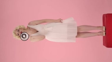 Marilyn Monroe karakterindeki kadın megafona konuşuyor. Stüdyoda pembe arka planda kırmızı kaide üzerinde duran muhteşem bir kadın. Dikey video. Satış, Kara Cuma. Boşluğu kopyala