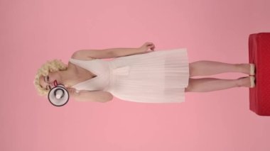Bir kadın megafona konuşur ve işaret eder, davet eder. Marilyn Monroe 'nun görüntüsündeki kadın pembe arka plandaki kırmızı kaidede dikiliyor. Dikey video