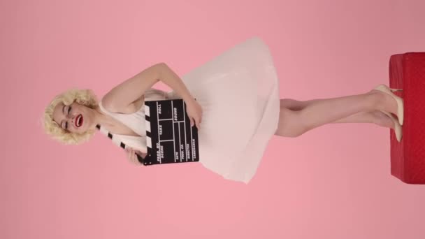 マリリン モンローがクラッパーボードを持っている姿を見た女性 風で発達する白いドレスの裾を保持し ピンクの背景にあるスタジオでペダルに立っている間にエアキスを送る女性 バーティカル — ストック動画
