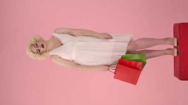 Alışveriş yorucu olabilir. Alnından ter silen alışveriş torbalı genç bir kadın. Marilyn Monroe 'nun görüntüsü stüdyoda pembe arka planda kaidede dimdik duran bir kadın. Dikey video