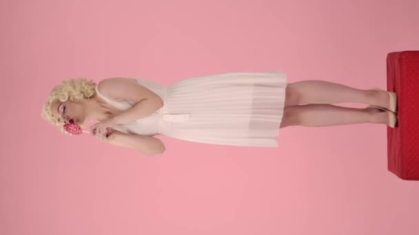 女性はカラフルなロリポップを保持し キスし 喜んでそれをライセンスします ピンクのバックグラウンドでスタジオのペデスタルに完全な高さに立っているマリリン モンローのイメージの女性 バーティカルビデオ スイートフードコンセプト — ストック動画