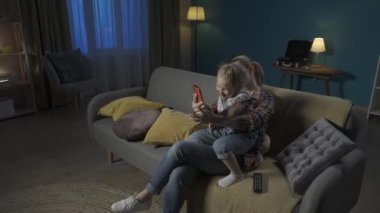 Mutlu anne ve kız, evdeki oturma odasında kanepede otururken akıllı telefondan selfie çekiyorlar. Sevimli küçük kız annesinin kollarında oturuyor ve komik suratlar yapıyor HDR BT2020 HLG Materyal