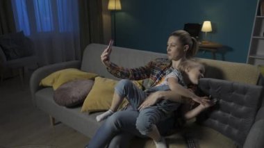 Mutlu anne ve kız, evdeki oturma odasında kanepede otururken akıllı telefondan selfie çekiyorlar. Sevimli küçük kız annesine sevgiyle sarılıyor ve annesi de kızını öpüyor HDR BT2020 HLG Malzeme