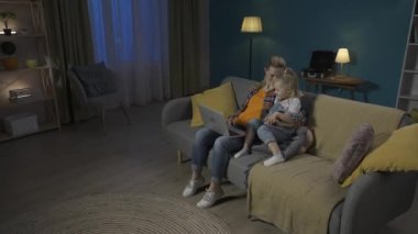 Mutlu, genç bir anne ya da dadı, genç kızıyla koltukta oturup dizüstü bilgisayarda video ve çizgi film izliyor. Anne ve kızı akşamları HDR BT2020 HLG 'de birlikte vakit geçirirler.